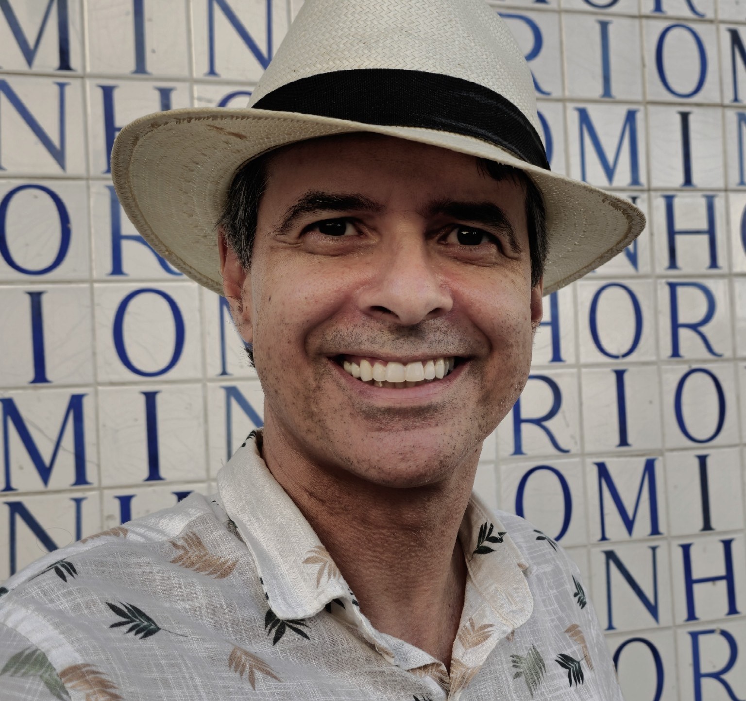Retrato de homem branco de chapéu, olhos castanhos escuros meio caidinhos, sorriso no rosto e camisa clara, com pequenos detalhes de folhas verdes e ocres-douradas. Ao fundo, azulejos com letras em azul.